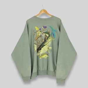 May include: Sage green sweatshirt featuring a colorful graphic of various fish species. The design includes yellow, blue, and green fish, with detailed illustrations. The sweatshirt is hanging on a wooden hanger, showcasing its relaxed fit and vintage style.