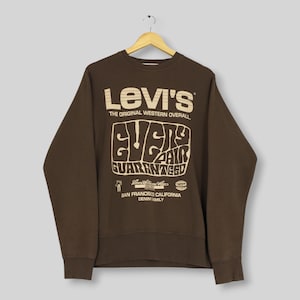 Könnte beinhalten: Ein dunkelbraunes Langarm-Sweatshirt mit Rundhalsausschnitt und einem großen cremefarbenen Grafikdruck auf der Vorderseite. Der Text lautet Levi's The Original Western Overall, Every Pair Guaranteed, Levi Strauss & Co, San Francisco California Denim Family.