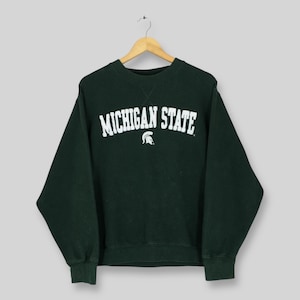 Puede incluir: Sudadera verde oscuro con el texto blanco "MICHIGAN STATE" en forma de arco. Un logotipo blanco de casco espartano está centrado debajo. La sudadera tiene cuello redondo y mangas largas.