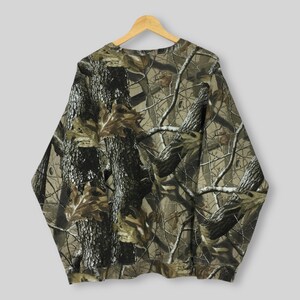 May include: A long-sleeved sweatshirt with a camouflage pattern. The design features a blend of brown, green, and tan hues, mimicking a forest environment. The fabric is a soft material, and the sweatshirt is hanging on a wooden hanger.