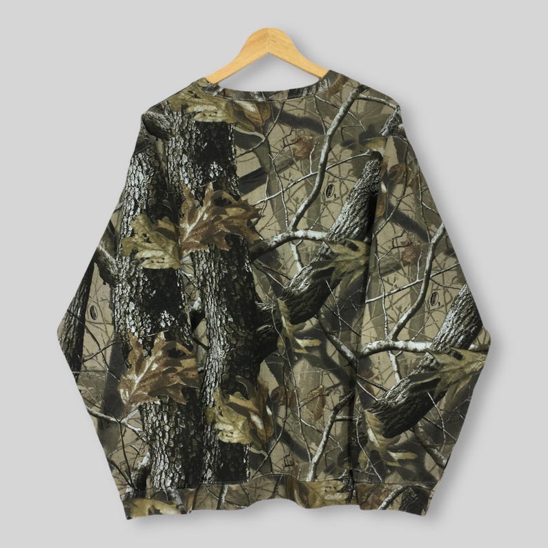May include: A long-sleeved sweatshirt with a camouflage pattern. The design features a blend of brown, green, and tan hues, mimicking a forest environment. The fabric is a soft material, and the sweatshirt is hanging on a wooden hanger.