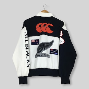 Puede incluir: Una camiseta de rugby de manga larga con un diseño de bloques de color en paneles negros y blancos. La parte trasera muestra un gran logotipo rojo de Canterbury, el texto ALL BLACKS, una bandera de Nueva Zelanda, un helecho y parches.