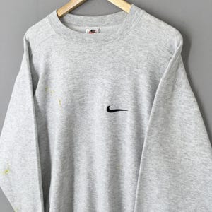 May include: A light grey Nike sweatshirt featuring a black Nike swoosh logo on the chest. The sweatshirt has a crew neck and long sleeves. The Nike logo is also visible on the upper chest. The sweatshirt is hanging on a wooden hanger.