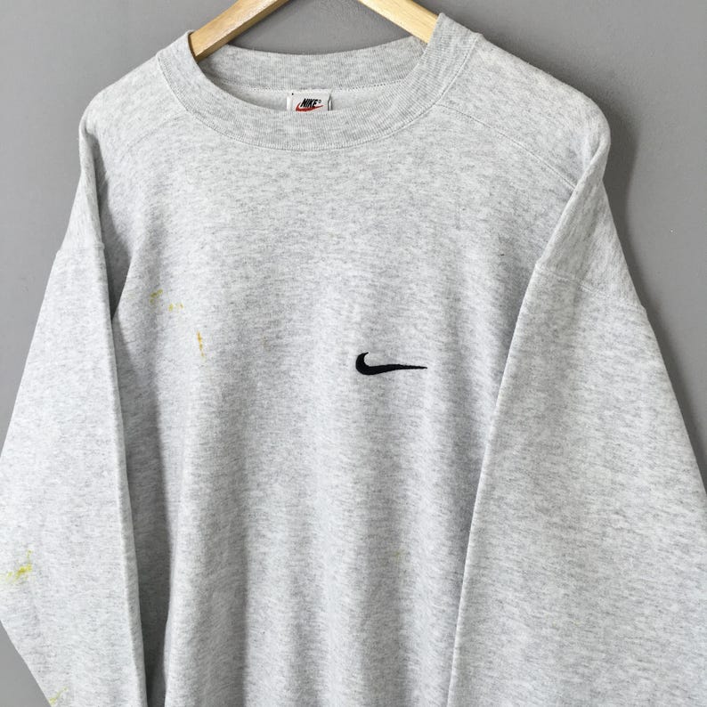 May include: A light grey Nike sweatshirt featuring a black Nike swoosh logo on the chest. The sweatshirt has a crew neck and long sleeves. The Nike logo is also visible on the upper chest. The sweatshirt is hanging on a wooden hanger.
