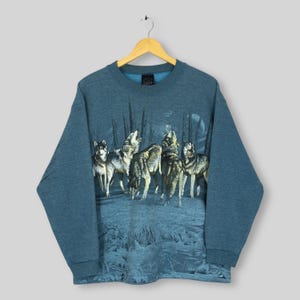 May include: A blue sweatshirt featuring a graphic of wolves howling at the moon. The wolves are depicted in a realistic style, with a forest and moon in the background. The sweatshirt has a crew neck and long sleeves.
