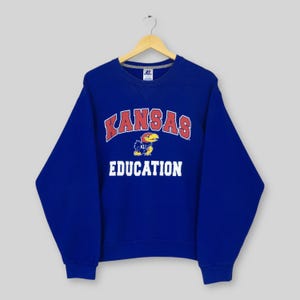 Pode incluir: Um moletom azul royal com a palavra "KANSAS" em letras vermelhas e brancas arqueadas. Abaixo, um mascote Jayhawk de desenho animado e a palavra "EDUCATION" em letras brancas. O moletom está pendurado em um cabide de madeira.