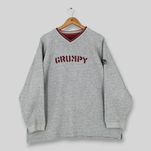 May include: A grey sweatshirt with a V-neck and long sleeves. The word "GRUMPY" is embroidered in maroon across the chest. The neckline has a maroon and navy blue trim. A small emblem is on the left sleeve.