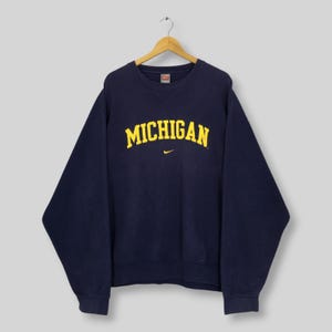 May include: A navy blue crewneck sweatshirt featuring the word MICHIGAN printed in yellow arched lettering across the chest. A small yellow Nike swoosh logo is positioned directly below the text. The garment is displayed on a wooden hanger.