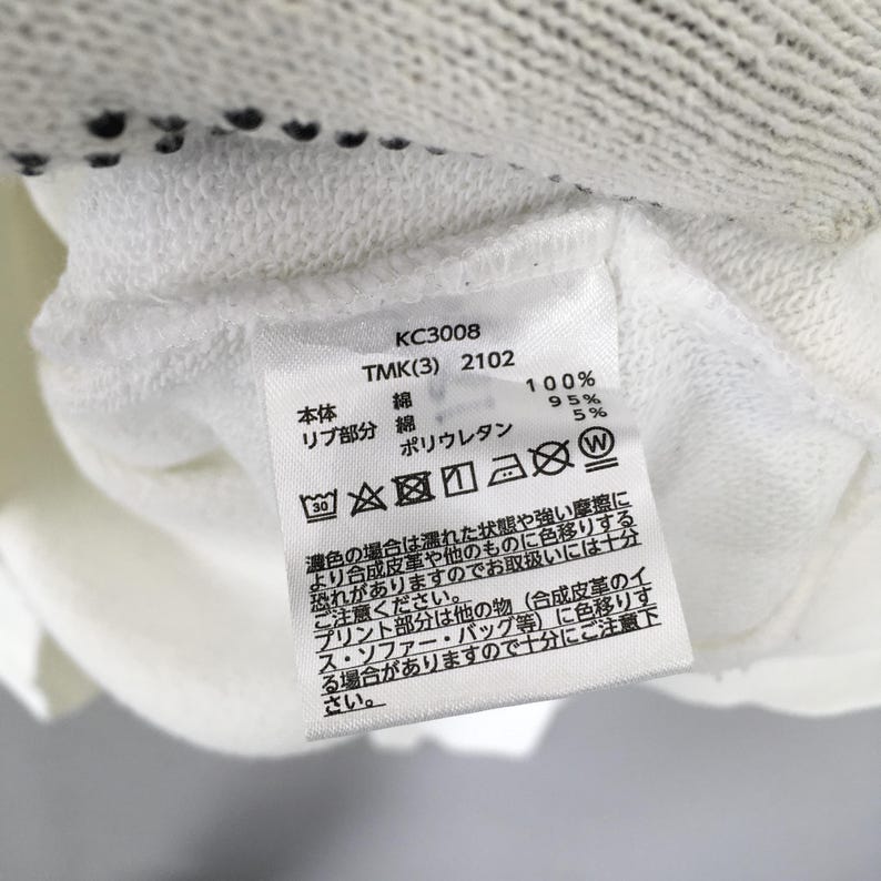 May include: A white fabric garment label featuring black text including KC3008, TMK(3) 2102, and material composition details of 95% cotton and 5% polyurethane. The label displays various laundry care symbols and Japanese text instructions for garment maintenance.