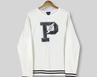 May include: White long-sleeved sweatshirt with a large black letter "P" on the front, featuring a winged emblem. The bottom and cuffs have black and white striped detailing. The sweatshirt is hanging on a wooden hanger.