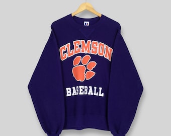 May include: A navy blue sweatshirt with the word "CLEMSON" in orange block letters above an orange paw print and the word "BASEBALL" in white block letters. The sweatshirt is hanging on a wooden hanger.