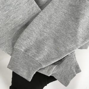 Puede incluir: Primer plano de una manga de sudadera gris con pu&ntilde;o acanalado. La tela es gris jaspeado y el pu&ntilde;o es de un tono gris m&aacute;s oscuro. La manga est&aacute; doblada sobre otra manga, con un vistazo de tela negra debajo.