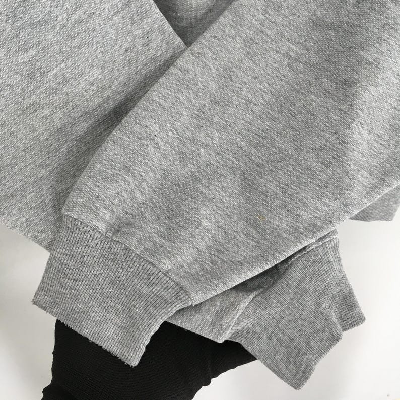 Puede incluir: Primer plano de una manga de sudadera gris con pu&ntilde;o acanalado. La tela es gris jaspeado y el pu&ntilde;o es de un tono gris m&aacute;s oscuro. La manga est&aacute; doblada sobre otra manga, con un vistazo de tela negra debajo.