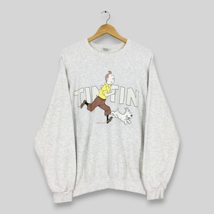 Peut inclure: Un sweat-shirt gris clair avec un dessin de Tintin courant avec Milou. Le mot "TINTIN" est imprim&eacute; en grandes lettres sur la poitrine. Les personnages de dessins anim&eacute;s sont en jaune, marron et blanc.
