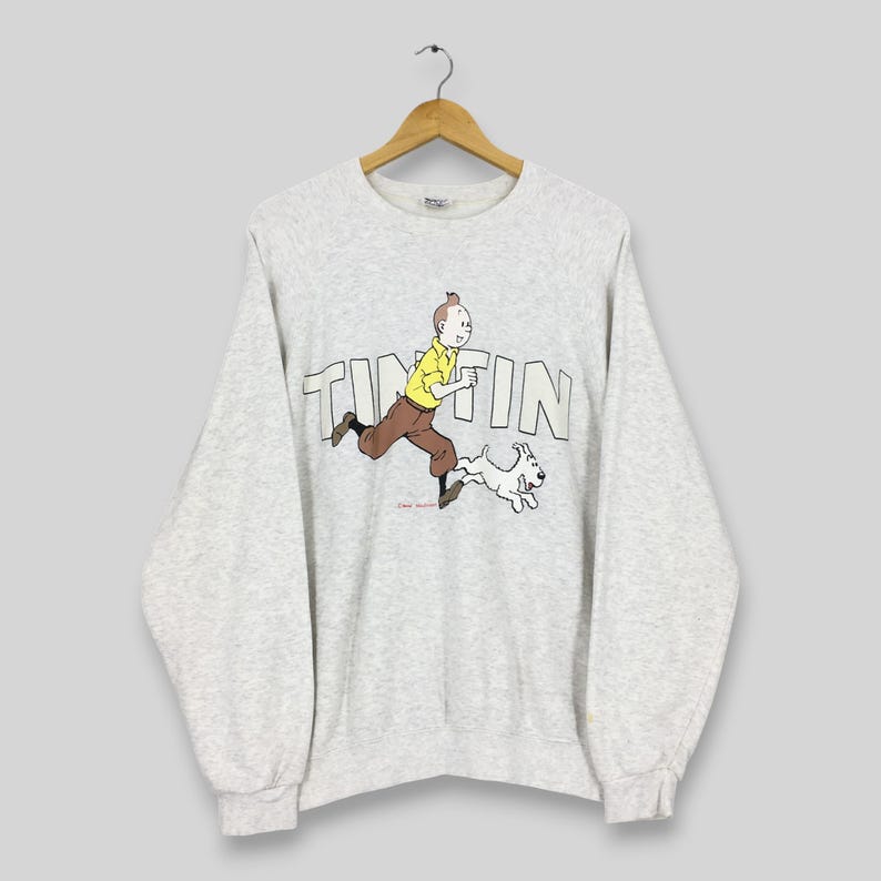 Peut inclure: Un sweat-shirt gris clair avec un dessin de Tintin courant avec Milou. Le mot "TINTIN" est imprim&eacute; en grandes lettres sur la poitrine. Les personnages de dessins anim&eacute;s sont en jaune, marron et blanc.