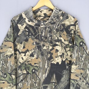 May include: A long-sleeved camouflage shirt with a cowl neck. The shirt features a pattern of brown, green, and gray leaves and branches. The shirt is hanging on a wooden hanger against a light gray background.