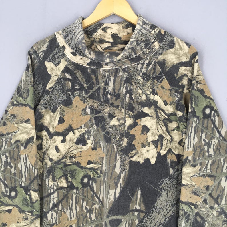 May include: A long-sleeved camouflage shirt with a cowl neck. The shirt features a pattern of brown, green, and gray leaves and branches. The shirt is hanging on a wooden hanger against a light gray background.