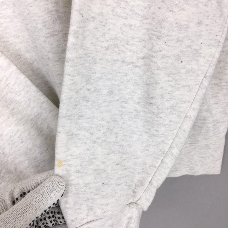 Peut inclure: Gros plan sur une manche de sweat-shirt gris clair. Le tissu a une texture mouchet&eacute;e avec quelques petits points sombres et une tache jaune. La manche est pli&eacute;e, r&eacute;v&eacute;lant la couture et le poignet.