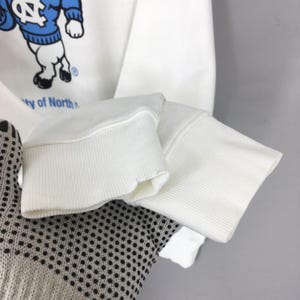 May include: A white long-sleeve sweatshirt featuring a graphic of a mascot wearing a blue jumper with a white NC logo. The graphic includes the text University of North Carolina. The sweatshirt has ribbed white cuffs at the end of the sleeves.