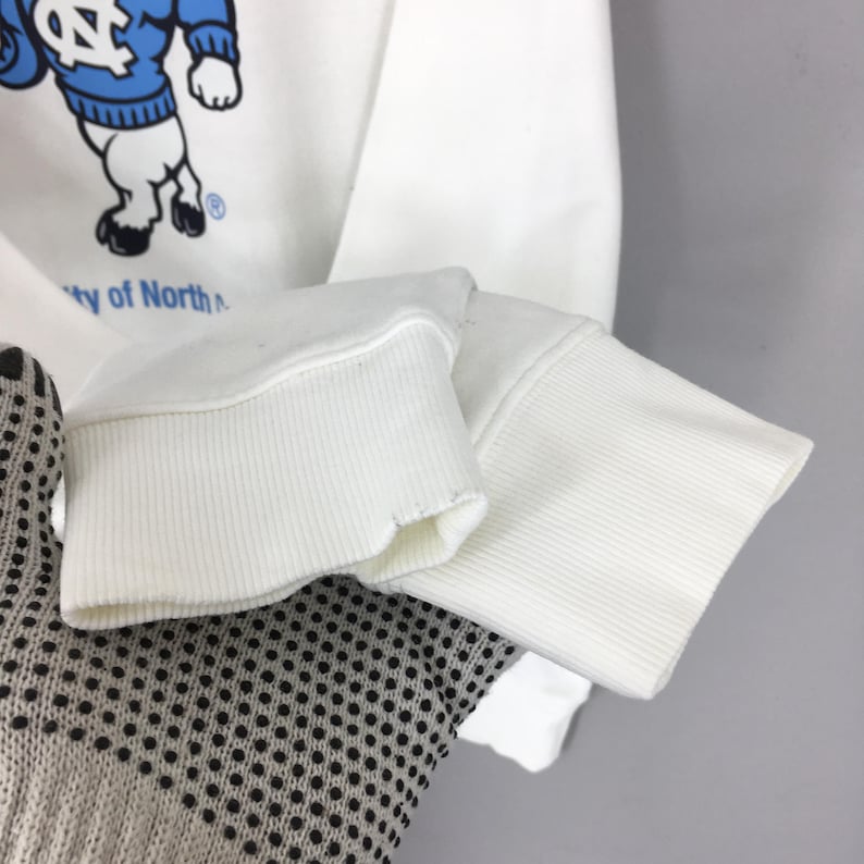 May include: A white long-sleeve sweatshirt featuring a graphic of a mascot wearing a blue jumper with a white NC logo. The graphic includes the text University of North Carolina. The sweatshirt has ribbed white cuffs at the end of the sleeves.