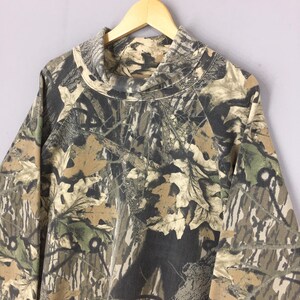 May include: A long-sleeved camouflage pattern shirt with a high, cowl neck. The shirt features a blend of brown, green, and gray tones, mimicking a forest environment. The fabric appears to be a medium-weight material, suitable for cooler weather.