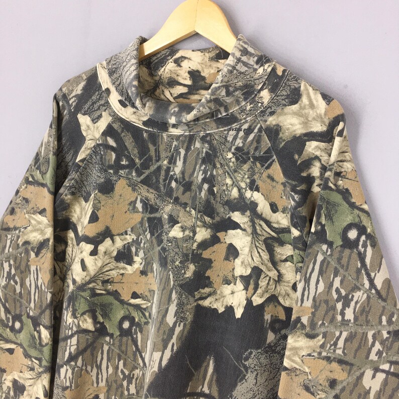 May include: A long-sleeved camouflage pattern shirt with a high, cowl neck. The shirt features a blend of brown, green, and gray tones, mimicking a forest environment. The fabric appears to be a medium-weight material, suitable for cooler weather.