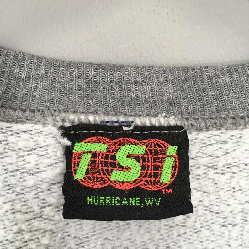 Puede incluir: Primer plano de un cuello de sudadera gris con una etiqueta negra. La etiqueta presenta las letras verde ne&oacute;n "TSI" sobre tres dise&ntilde;os de globo rojos y negros. El texto "HURRICANE, WV" est&aacute; debajo.