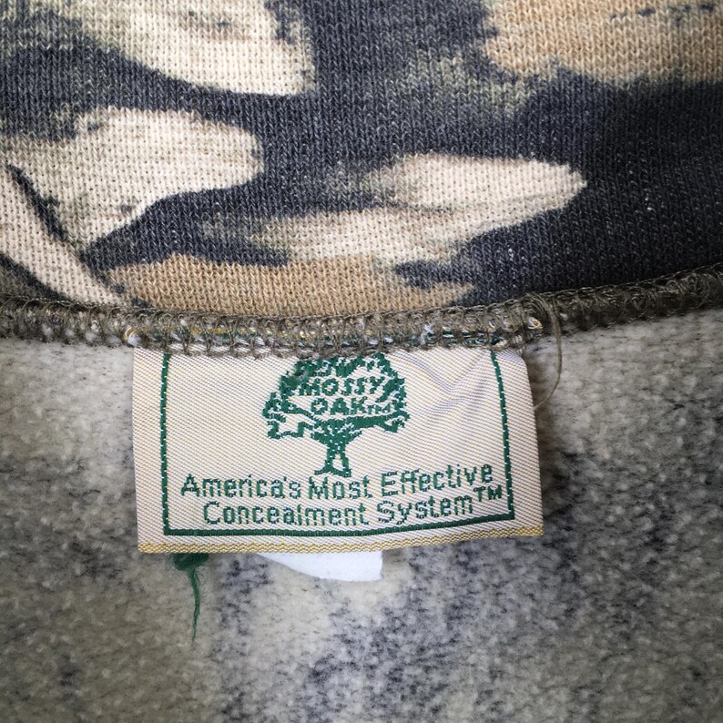 May include: Close-up of a camouflage pattern fabric with a tag. The tag features a green tree logo and the text "America's Most Effective Concealment System". The fabric has a mix of green, brown, and beige tones.