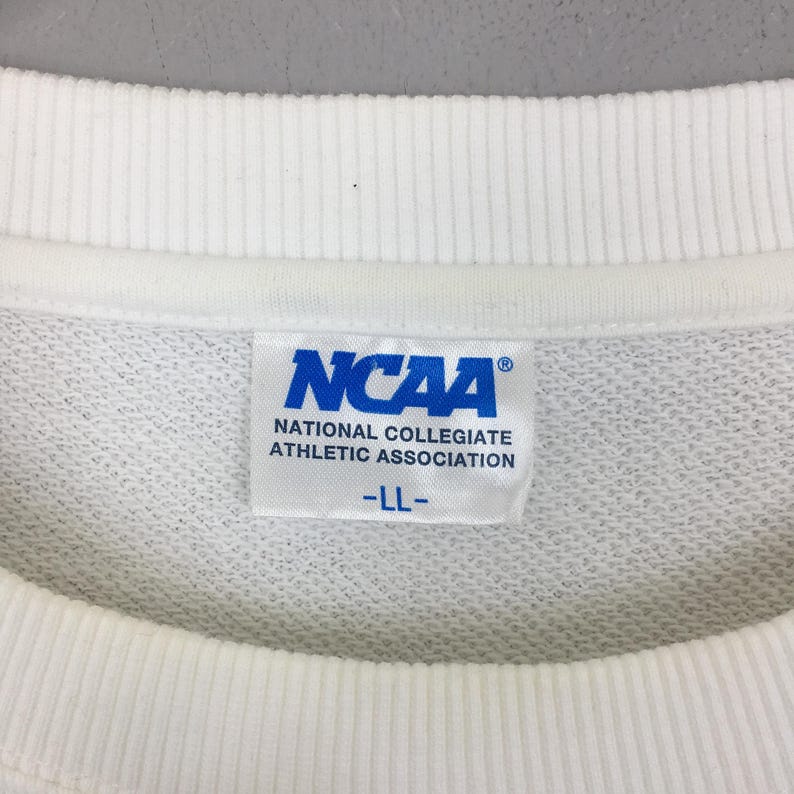 May include: A white crew neck sweatshirt featuring a white fabric tag on the inside collar. The tag displays the blue NCAA logo, the text National Collegiate Athletic Association, and the size LL printed in blue on a white background.