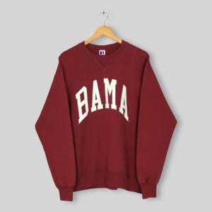 May include: Maroon sweatshirt with the word "BAMA" in white, arched lettering. The sweatshirt is hanging on a wooden hanger. The Russell Athletic logo is visible on the upper chest.
