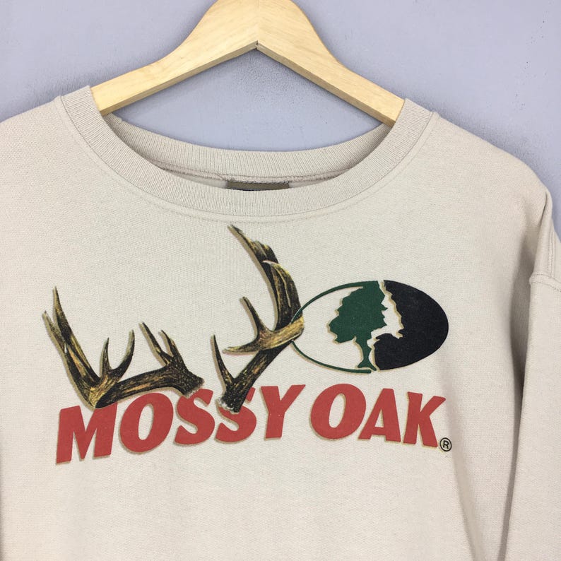 May include: Beige sweatshirt with the Mossy Oak logo. The logo features brown antlers, a green tree, and the words "Mossy Oak" in red. The sweatshirt is hanging on a wooden hanger.