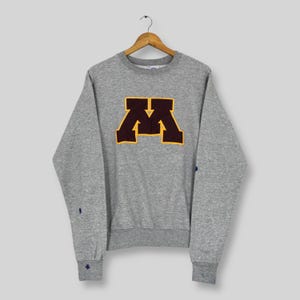 May include: A gray Champion sweatshirt with a large maroon and gold "M" logo on the front. The long-sleeved crew neck sweatshirt has a ribbed collar, cuffs, and hem. Small blue and navy details are visible on the sleeves.