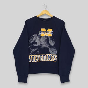 May include: A navy blue long-sleeve sweatshirt featuring a graphic of a wolverine in white and gray. Above the graphic is a yellow and blue block letter M with the text MICHIGAN. Below the graphic is the text WOLVERINES in yellow and blue.