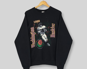 Op de afbeelding: Zwart sweatshirt met een afbeelding van een American footballspeler. De afbeelding bevat de tekst "1992 Washington Huskies Rose Bowl Tournament". Het ontwerp is paars, groen en wit. Het sweatshirt hangt aan een houten hanger.