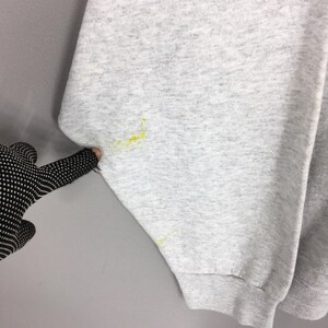May include: Close-up of a light grey sweatshirt sleeve with yellow stains. The sleeve is cuffed at the wrist. A gloved hand is pointing at the sleeve.