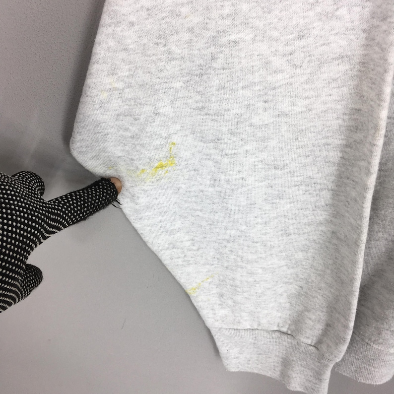 May include: Close-up of a light grey sweatshirt sleeve with yellow stains. The sleeve is cuffed at the wrist. A gloved hand is pointing at the sleeve.