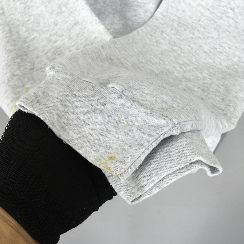 May include: Close-up of a grey sweatshirt sleeve with a black cuff. The grey fabric shows slight discolouration with yellow spots. The cuff is a solid black colour.