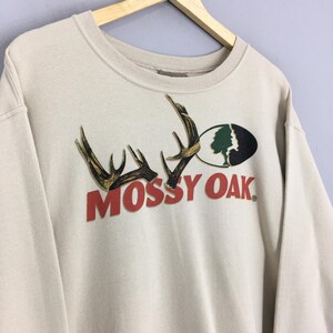 May include: A beige sweatshirt displaying the "Mossy Oak" logo in red. The logo incorporates antlers and a tree graphic. The sweatshirt is suspended on a wooden hanger.