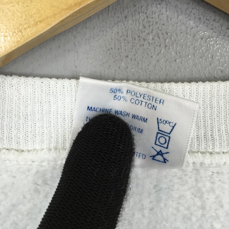 May include: Close-up of a white garment tag with text indicating it is 50% polyester and 50% cotton. The tag also includes washing instructions: machine wash warm at 50&deg;C. The garment is white.
