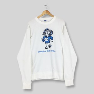 May include: A white long-sleeve crewneck sweatshirt featuring a graphic of the University of North Carolina mascot, a ram wearing a blue sweater with the letters UNC, and the text University of North Carolina printed in blue below the mascot.