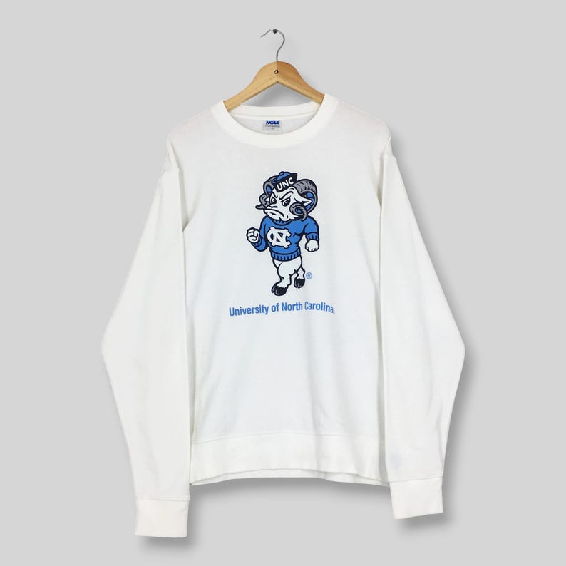 May include: A white long-sleeve crewneck sweatshirt featuring a graphic of the University of North Carolina mascot, a ram wearing a blue sweater with the letters UNC, and the text University of North Carolina printed in blue below the mascot.
