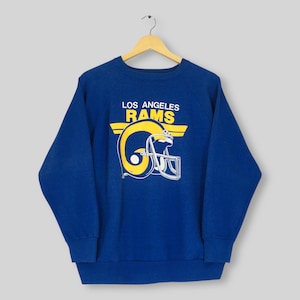 Puede incluir: Una sudadera azul con las palabras "LOS ANGELES RAMS" en blanco y amarillo sobre un gráfico de casco de fútbol amarillo y blanco. La sudadera está colgada de una percha de madera.