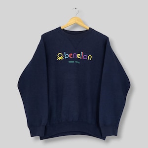 Puede incluir: Una sudadera de cuello redondo y manga larga en color azul marino con la palabra benetton bordada en letras multicolores en el pecho, y las palabras UNITED COLORS bordadas en letras verdes más pequeñas justo debajo del logotipo.