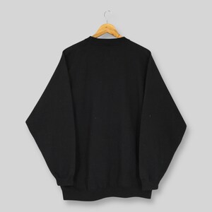 May include: A black crew neck sweatshirt hanging on a wooden hanger. The sweatshirt is a solid black color and has long sleeves. The garment is displayed against a light gray background.