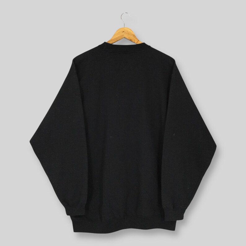 May include: A black crew neck sweatshirt hanging on a wooden hanger. The sweatshirt is a solid black color and has long sleeves. The garment is displayed against a light gray background.