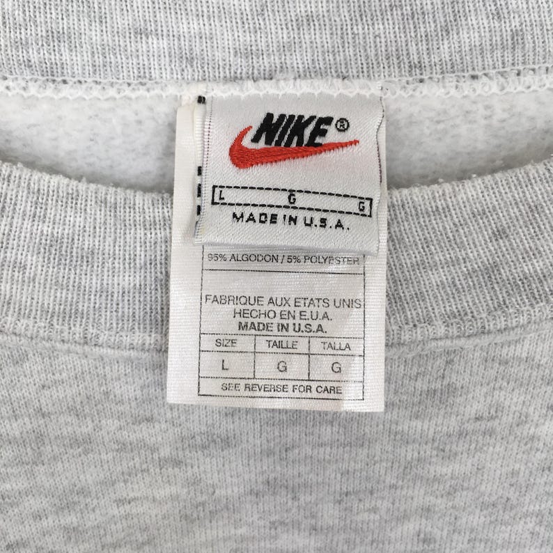 May include: Close-up of a grey sweatshirt with a Nike label. The label shows the Nike logo, text reading "MADE IN U.S.A.", and size details. The fabric composition is 95% cotton and 5% polyester.