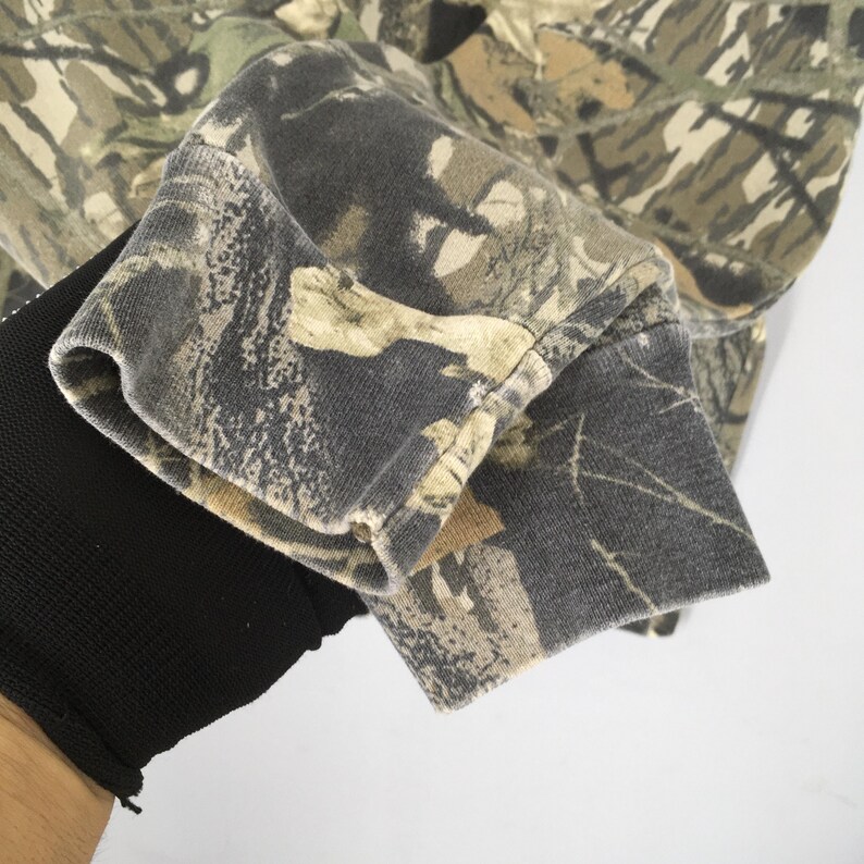 May include: Close-up of a camouflage-patterned long-sleeved shirt. The fabric features a blend of greens, browns, and grays, creating a woodland effect. The cuff of the sleeve is visible, showing the texture and design of the material. The shirt appears to be made of a soft, durable fabric.