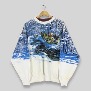 May include: White sweatshirt with a winter scene print. The design features a horse-drawn sleigh carrying people through a snowy forest. The sleeves and lower portion of the sweatshirt are white, while the upper portion has a blue and white winter landscape.