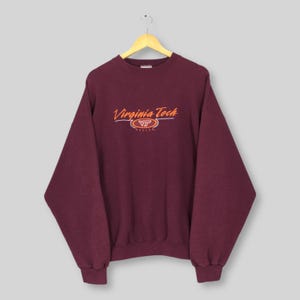 Op de afbeelding: Een bordeauxrode Virginia Tech-trui. De woorden "Virginia Tech" staan in oranje letters boven een oranje "VT" logo. De trui hangt aan een houten hanger tegen een lichte achtergrond.