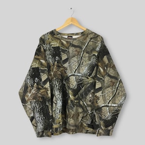 May include: A long-sleeved camouflage sweatshirt with a crew neck, hanging on a wooden hanger. The pattern features a mix of brown, green, and tan, resembling a forest scene. The sweatshirt is made of a soft material.
